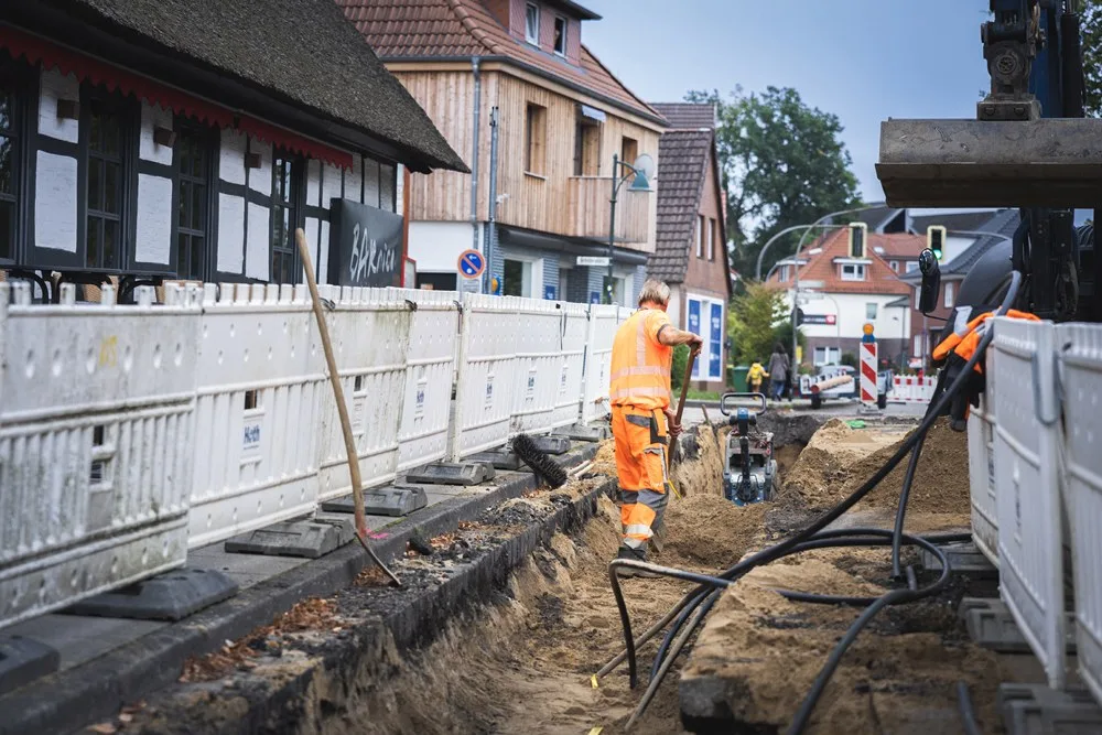 Kabelgraben entlang eines Bauzauns, ein Arbeiter in Warnkleidung, schwarze Kabel liegen im Graben